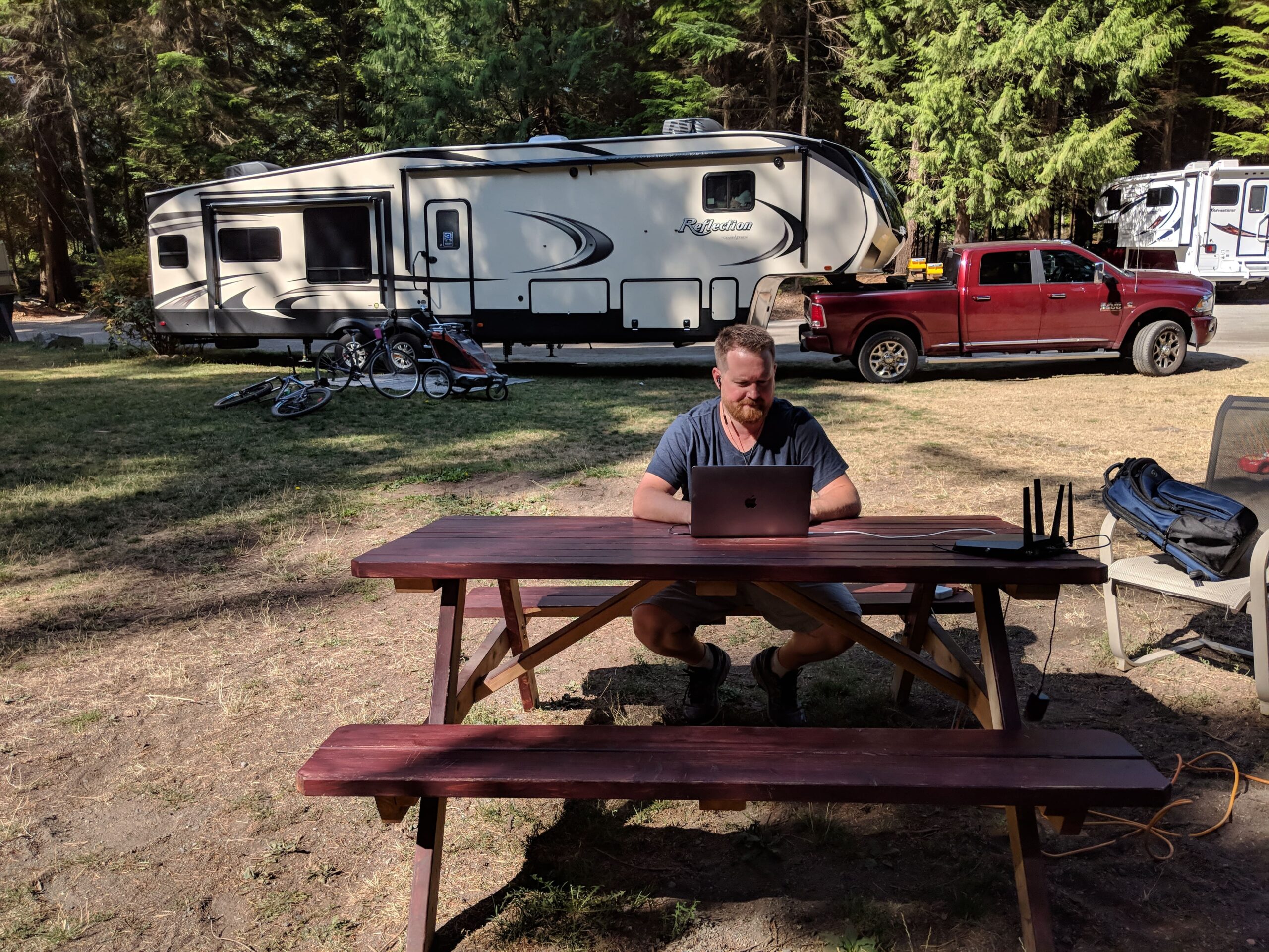 A man working on a laptop at a picnic table, with an RV and trees in the background.